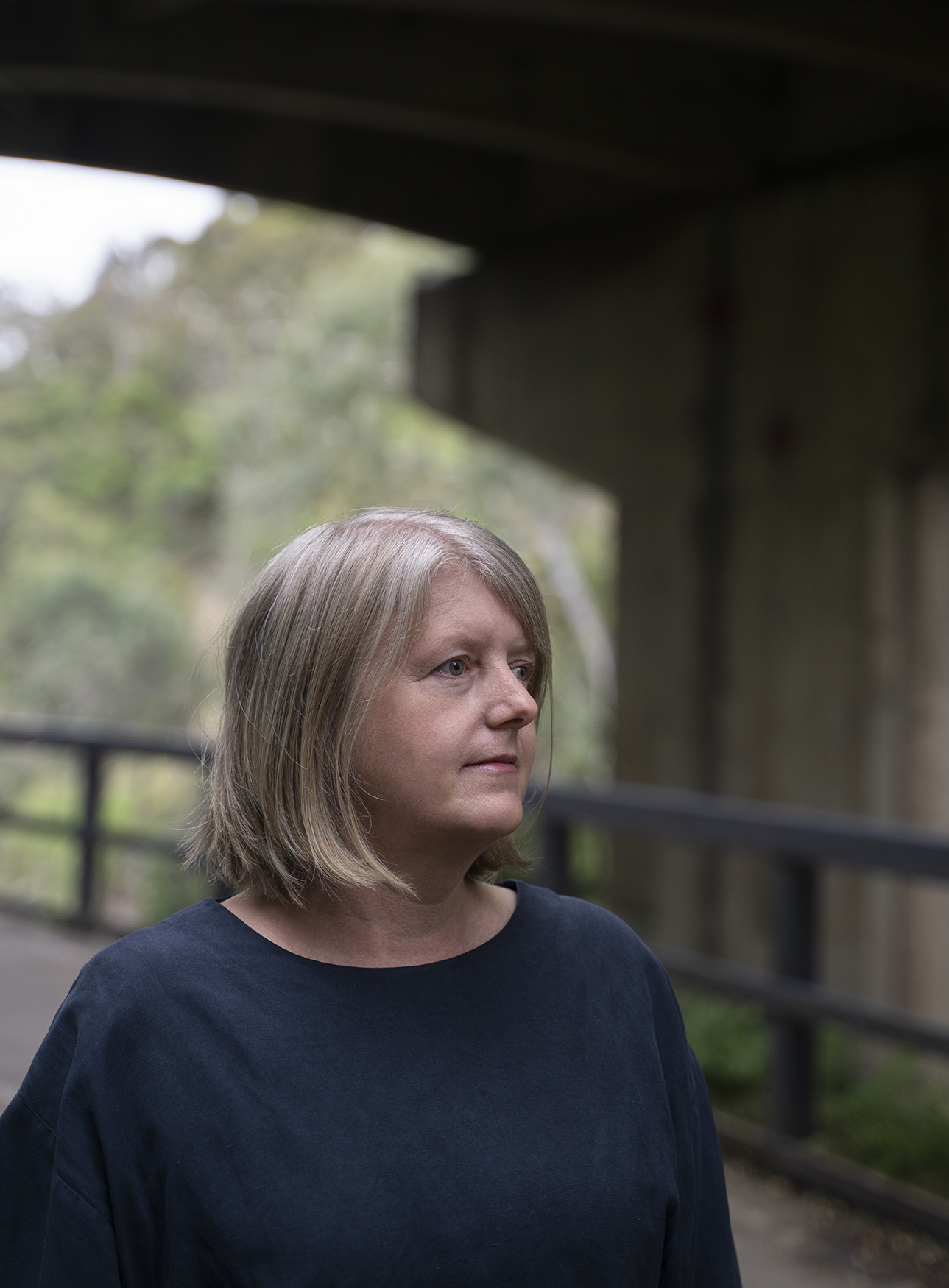 A blond middle-aged woman in a blue top looks melancholy as she stands under a bridge.